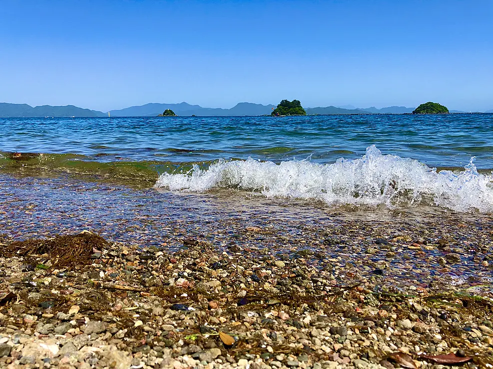 熊本県津奈木町にある三ツ島海水浴場から、沖に浮かぶ3つの島（竹島、木島、沖島）を望む風景。手前には小石の海岸と穏やかな波が打ち寄せ、晴れた青空が広がっている。