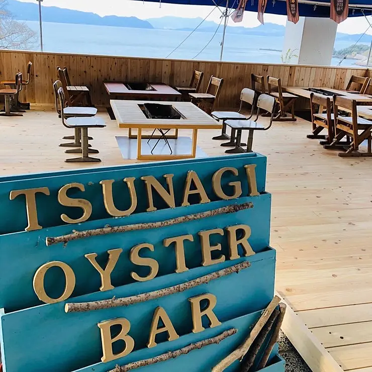 「TSUNAGI OYSTER BAR」の看板がかかった牡蠣小屋の木製テーブルと椅子がある内装