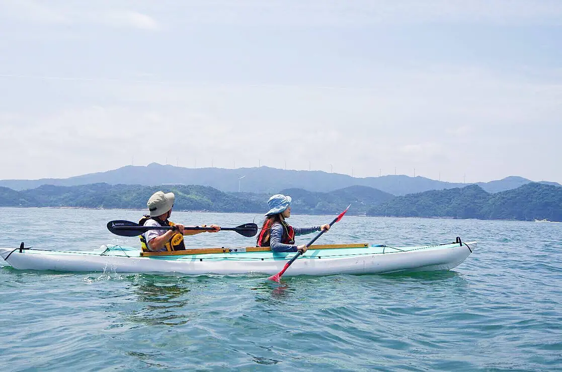 海上で二人乗りのカヤックに乗る人　奥には穏やかな山並み