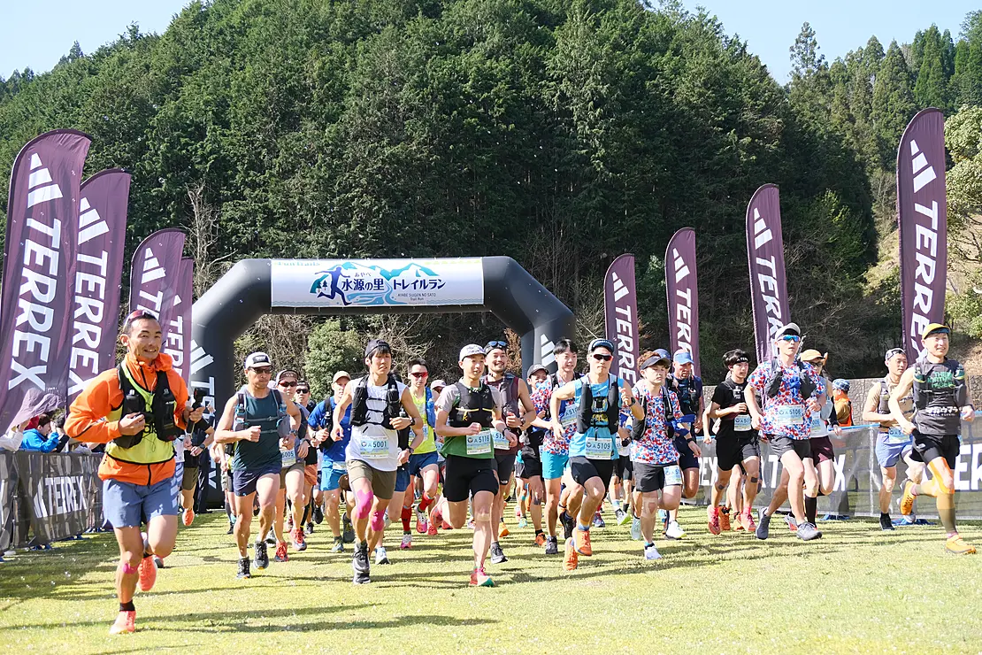 緑豊かな山々を背景に、トレイルランナーたちがスタート地点のアーチをくぐり抜けている様子