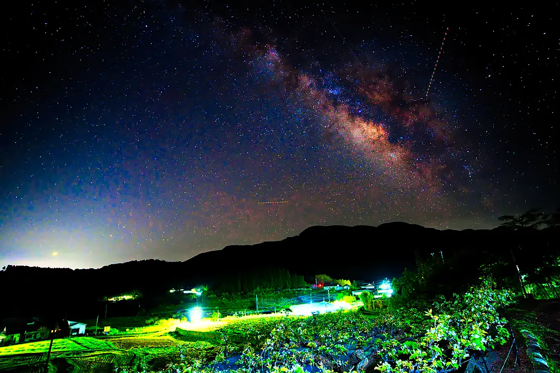 毎床地区の梨園上空に広がる天の川の夜景。暗闇の中、星空と畑の様子。