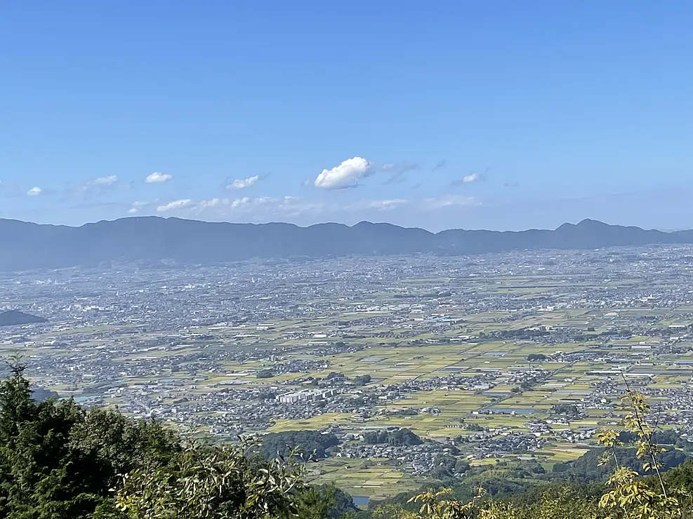 山の上から見下ろした奈良盆地　手前には緑の木々、その奥には田畑と民家が広がり、遠くには連なった山並みが横たわっている
