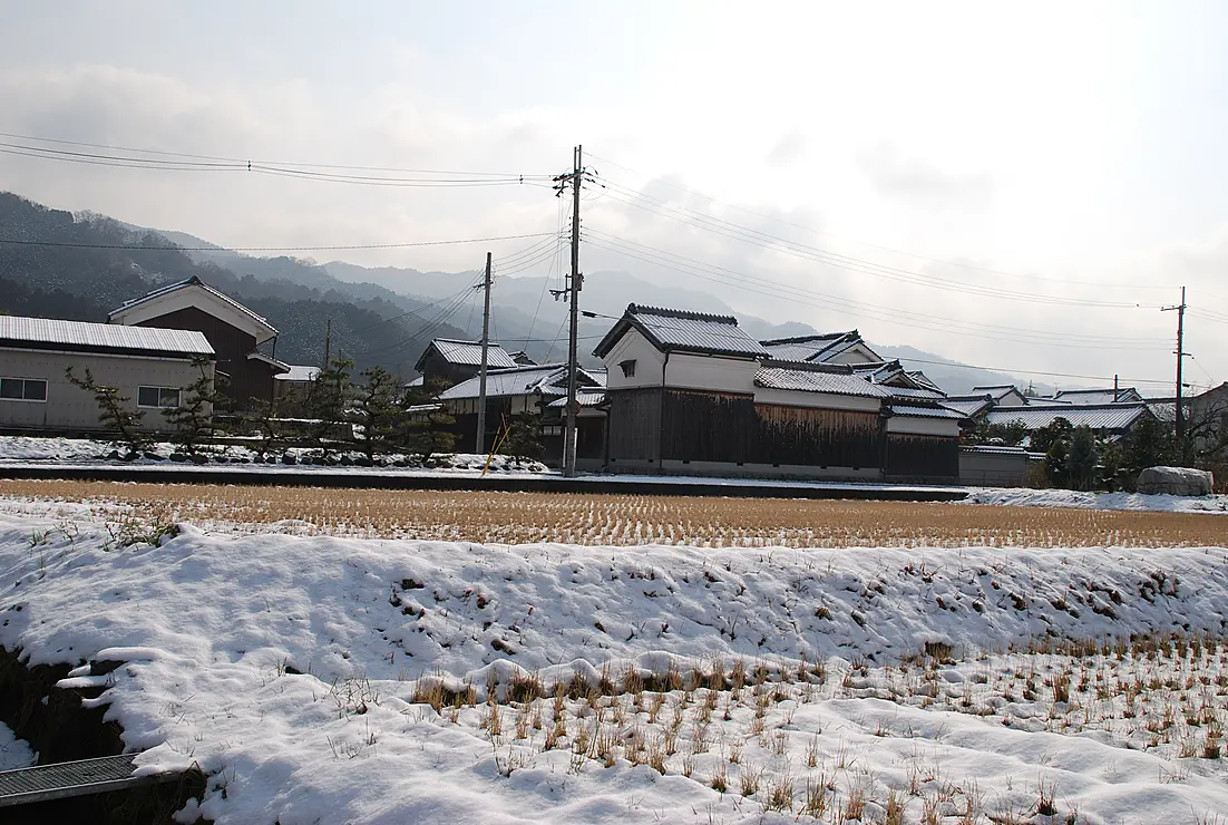 雪が薄く積もった田んぼと、その奥に伝統的な家屋。背景には山々が霞んで見える