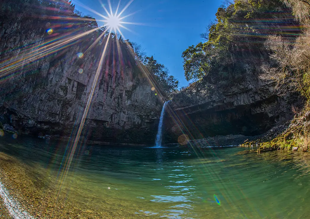 洞窟のような岩壁から水が流れ落ちる「うのこの滝」。水しぶきと木々に明るい日差しが差し込んでいる。