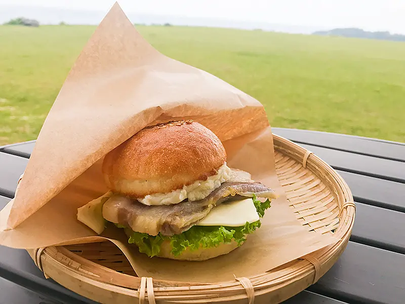 籐のトレイに乗せられた「塩豚バーガー」が写っています。バンズには奄美の伝統料理である塩豚、オーガニック野菜、マッシュポテト、チーズが挟まれています。