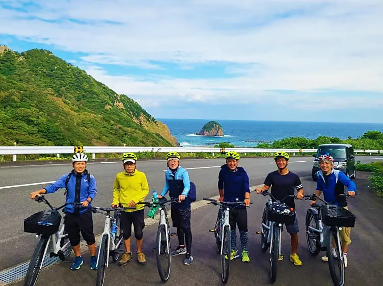 奄美大島の宮古崎で、自転車に乗った6人の男女が海と緑豊かな丘を背景にポーズをとっている様子