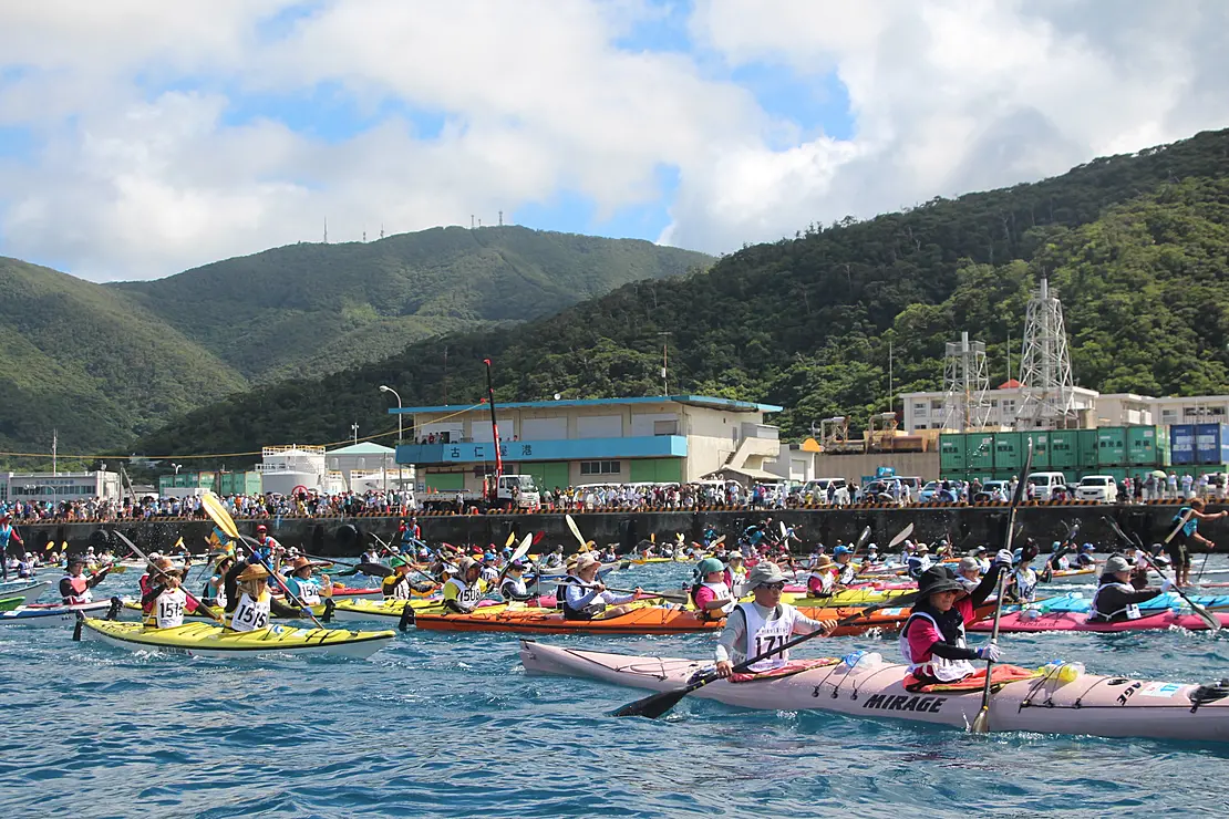 多くの参加者が乗った色とりどりのシーカヤックが海上に浮かんでおり、皆が一方向に向かっています。背景には緑豊かな山々が広がり、海岸線にはいくつかの建物が見えます。