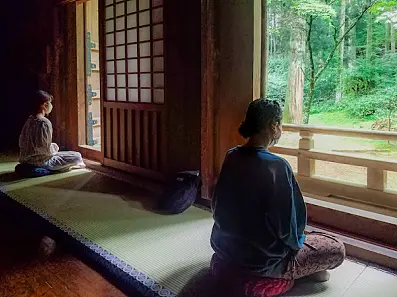 伝統的木造建築の窓際に敷かれた畳の上で、外を眺めながら座禅を組む人