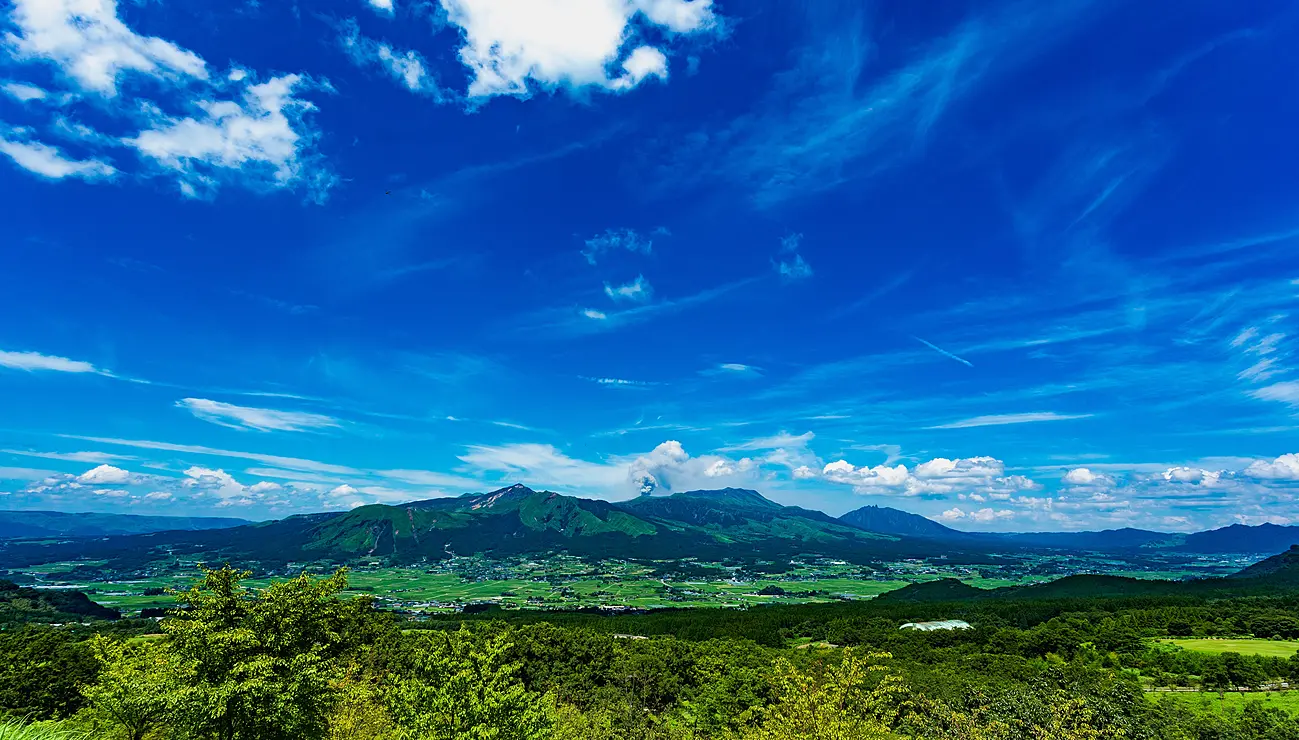 阿蘇五岳と南外輪山を一望する雄大な風景。青空と緑豊かな草原が広がる。