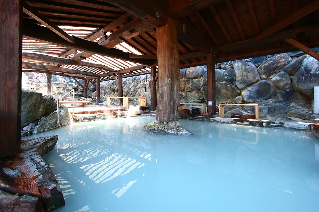 木造の屋根と岩壁に囲まれた、乳白色の青い湯が満ちた温泉の露天風呂