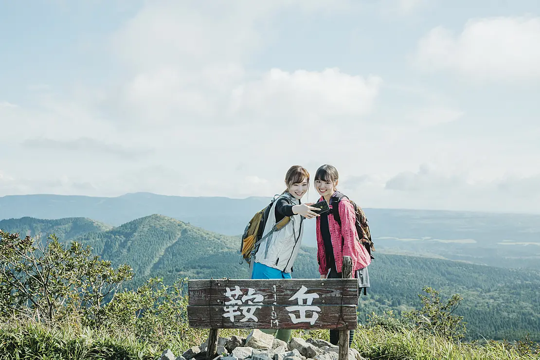 山々と湖を背景に鞍岳の看板前で写真を撮る二人