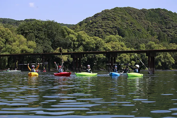 四万十川にかかる沈下橋を背景に色とりどりのカヤックに乗った人達が楽しそうにパドルを空へ掲げています。