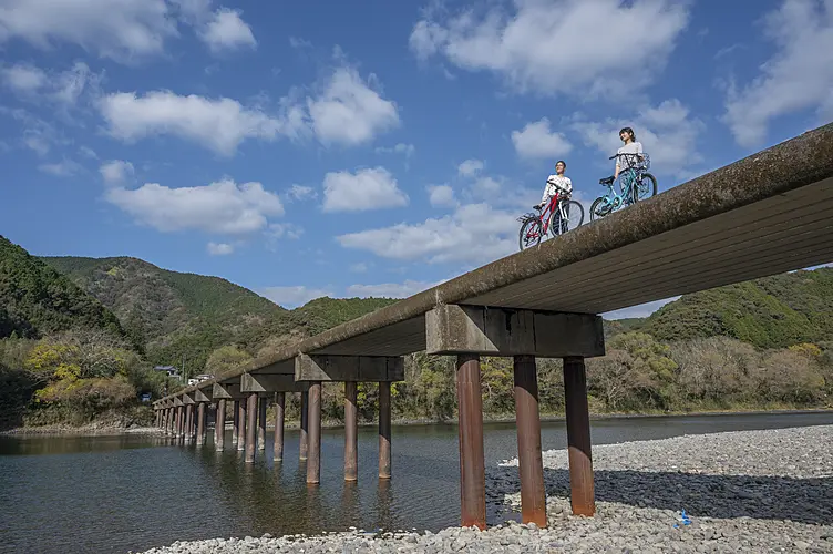 晴天のなか、四万十川に架かる沈下橋を自転車で女性2人が渡っている